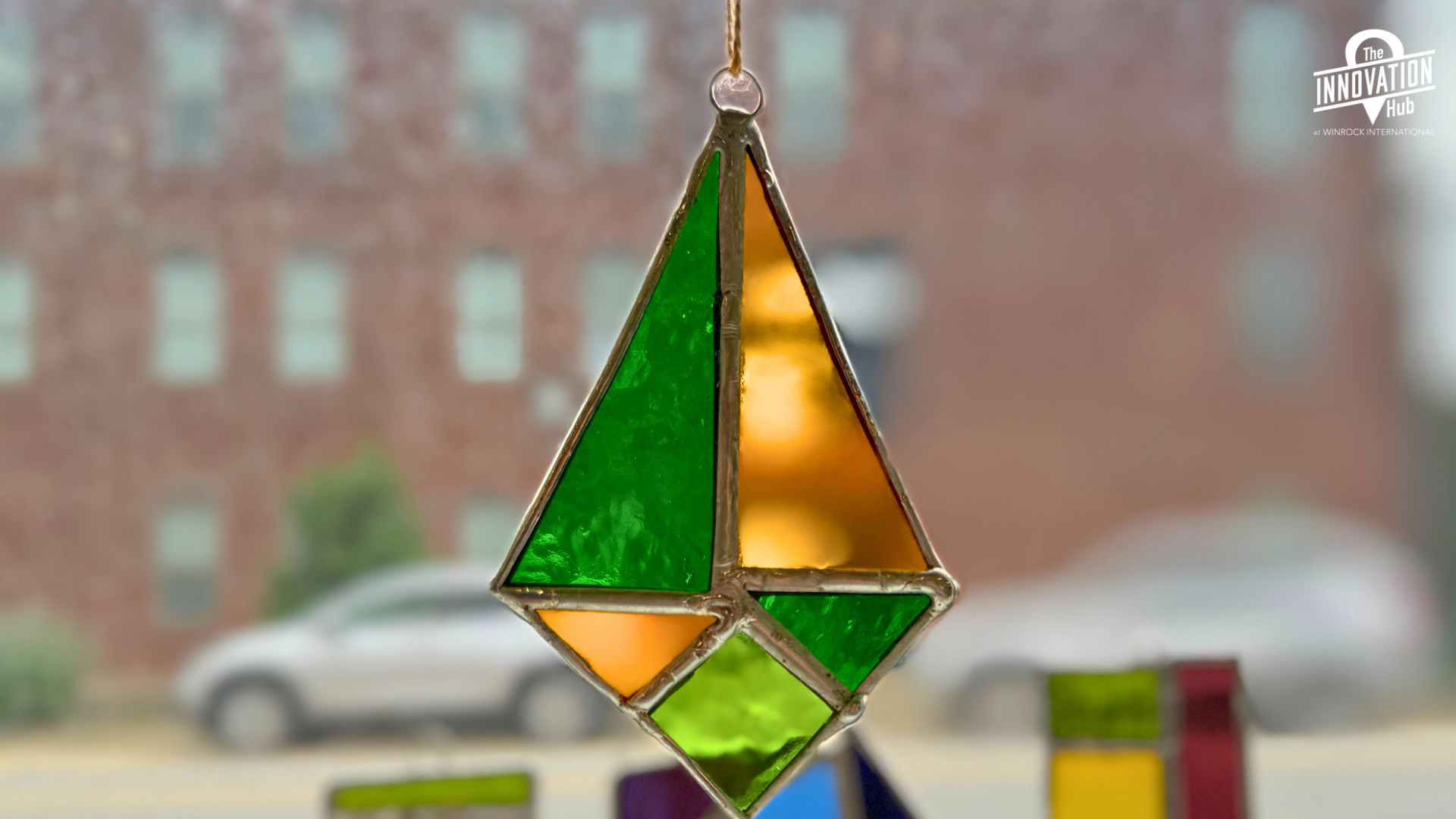 A stained glass sun catch in green and gold hangs in front of a window with other glass suncatchers in the background.