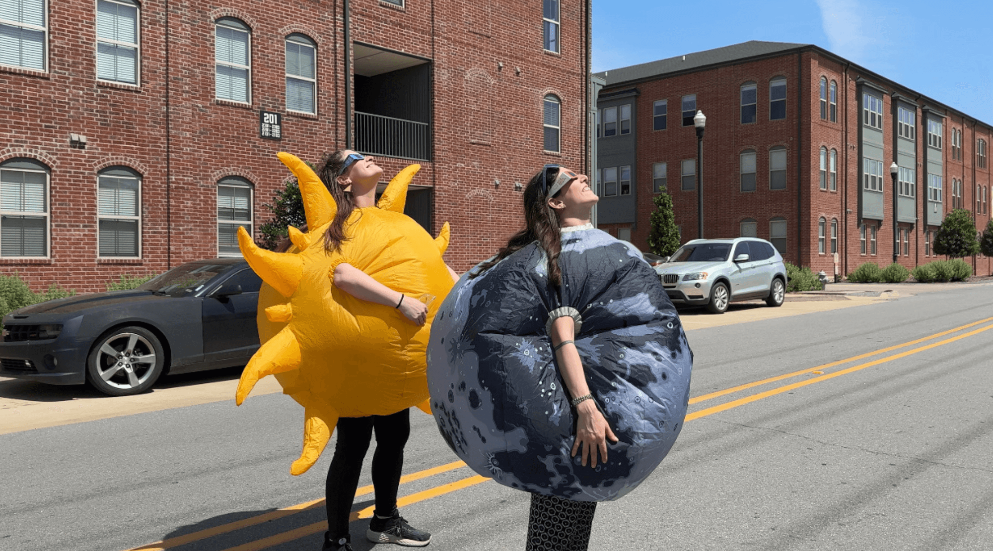 Two Hub staff members stare up at the sky while wearing blow up costumes.  One is wearing a yellow sun and the other is wearing a moon.  They wear solar eclipse glasses to protect their eyes.