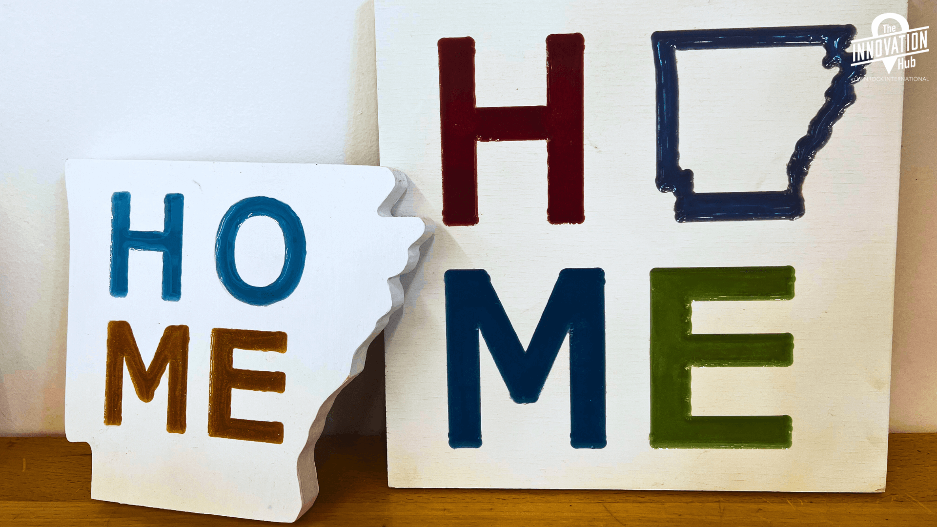 A photo of two different signs. The sign on the left is in the shape of Arkansas and says "HOME" in different color epoxies. The sign on the right is square and says "HOME" with the O as a shape of Arkansas and has colorful epoxies.