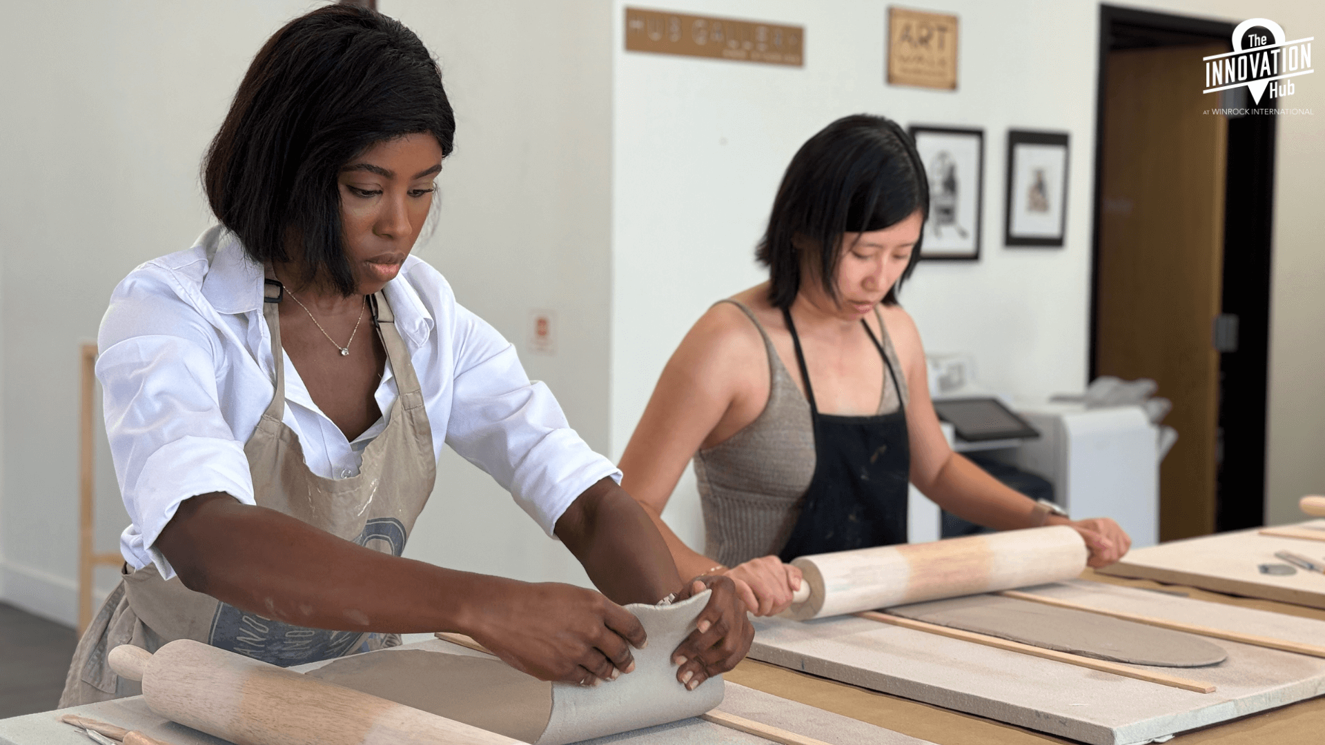 Two women working on canvas boards with wooden rollers to flatten out their clay.