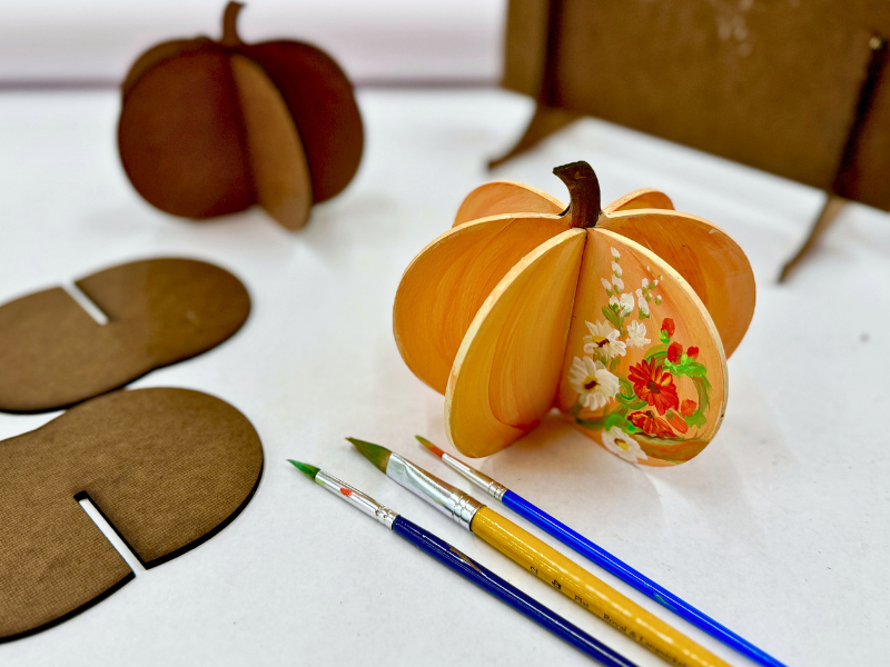 A laser cut pumpkin has been painted in pretty fall colors with a spray of fall flowers. In the background are recently cut panels that stack together to form the 3d shape.