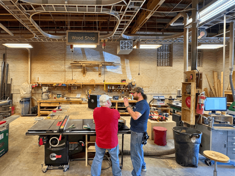 Adam, the Woodshop foreman, speaks with a guest in the woodshop next to a large table saw.