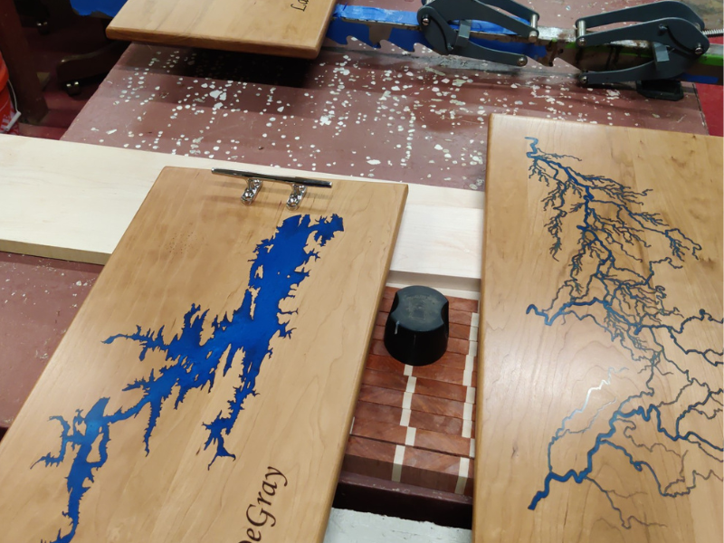 Several  wooden trays with deep blue epoxy dry on racks.  Each tray is a lovely walnut brown and in the middle is the topographical region of Lake Degray, filled in with blue.